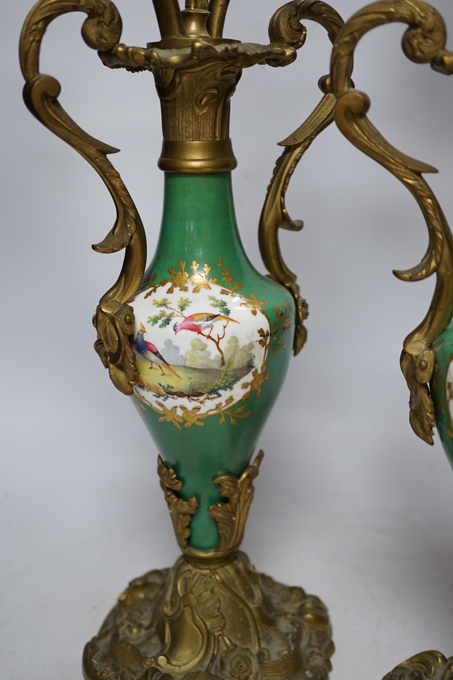 A pair of Sevres style porcelain and brass rococo-style two branch candelabra, hand painted with exotic birds, 50cm high